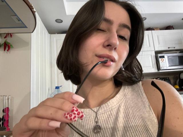 KamilaHarison with shoulder-length brown hair, pink manicured nails, sucking on a black straw, wearing a beige ribbed sleeve top, black strap visible, in a kitchen.
