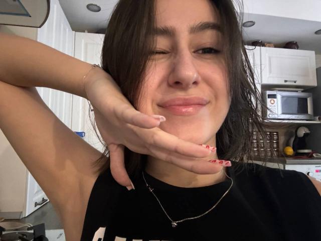 KamilaHarison with dirty brown hair, winking, black shirt, holding chin with red-nailed hand, silver necklace, subtle smile, in a kitchen. Her ass is not visible.