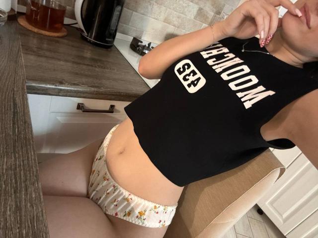 KamilaHarison in a black sleeveless top, showing her flat stomach and floral panties, with one hand near her mouth, sitting on a kitchen chair. Boobs not visible. Ass not visible.