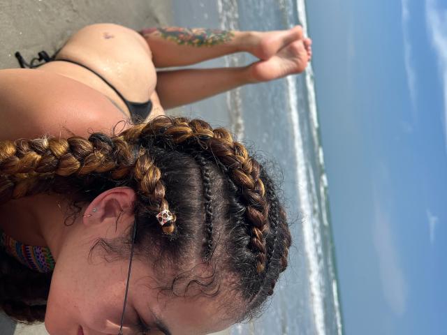 HellenK mit geflochtenem, braunem Haar, trägt einen schwarzen String-Bikini und liegt auf einer Betonfläche, mit dem Gesäß nach oben, die Beine im Wasser, hat ein Tattoo am rechten Arm und trägt einen Ohrstecker..