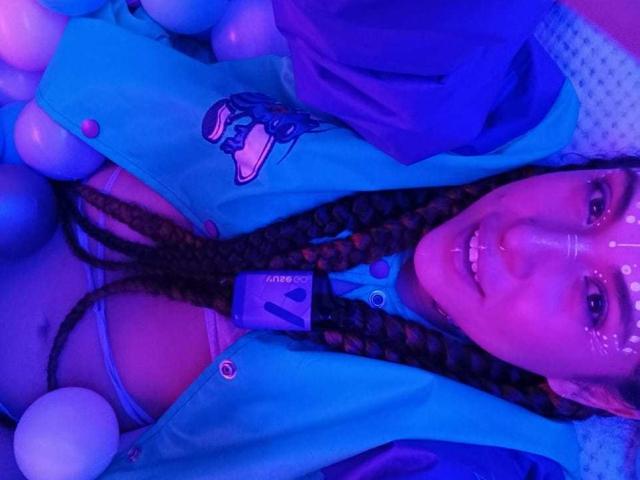 HellenK with braided hair, blue jacket, and blue bra, surrounded by balloons, has a facial mask and a smiling face, showing tits, in purple-blue lighting.