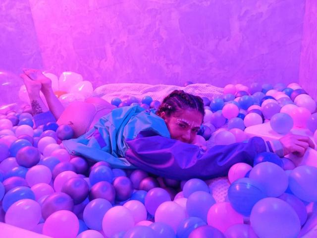 HellenK with braided hair, glasses, and blue jacket, laying in a pink and purple ball pit, arms and legs partially hidden, looking at camera with a relaxed expression.