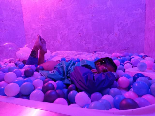 HellenK laying in a blue and white ball pit, wearing a blue hoodie, with legs up, pink and purple lighting, arms covering face, relaxed, in a colorful, bubbly environment.