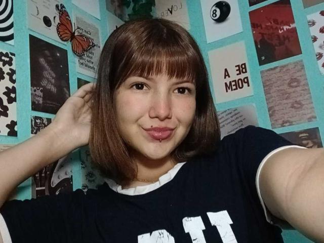 Caramelpearl with brown bob, pouty lips, and a septum piercing, wearing a black tee with white trim, hand in hair, smiling, teal wall of photos in background.