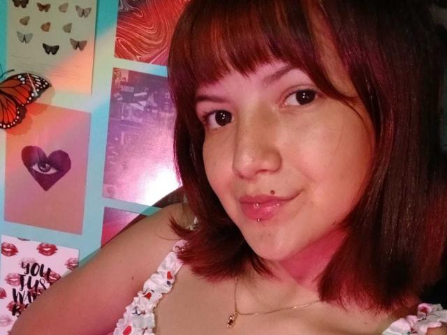 Caramelpearl with red-brown hair, freckles, and a lip piercing, wearing a white floral top, smiling at the camera. Her nose has a small beauty mark.