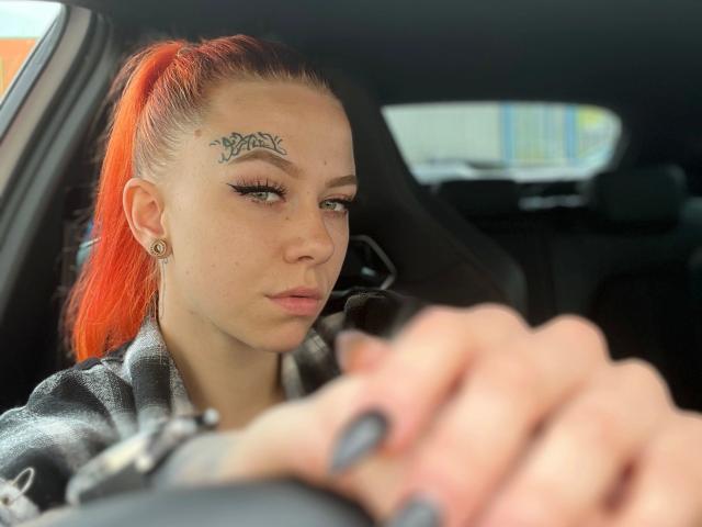 MeriJey – mit rotem Pferdeschwanz, Augenbrauen-Tattoo, schwarzem Eyeliner und Nasenring, beim Autofahren, Hände am Lenkrad, schwarze Nägel, ernster Blick, trägt eine graue Jacke, Innenraum eines Autos..