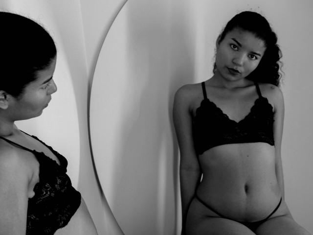 Foto in bianco e nero di SexLaddy con i capelli ricci, che indossa un reggiseno e uno slip di pizzo, in piedi e che si guarda allo specchio, mostrando i suoi piccoli seni e un leggero girovita..