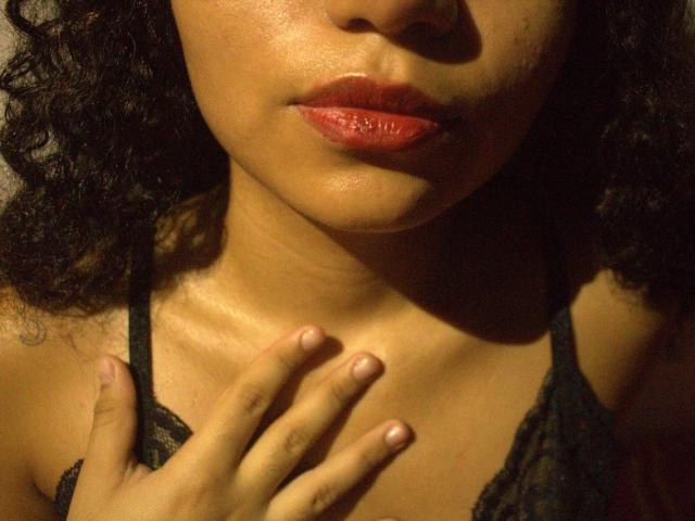 Das Gesicht einer Frau namens SexLaddy mit dunklen, lockigen Haaren, roten, glänzenden Lippen und brauner Haut, die ein schwarzes Spitzenoberteil trägt, mit einer Hand auf der Brust, weiche Beleuchtung, leicht glänzende Haut, warmer Ton..