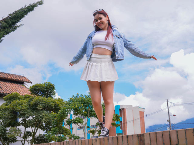 MiaPoe with red hair, blue jacket, white crop top, and white skirt, standing on a brick wall, arms out, smiling, wearing black sneakers, cloudy sky above.