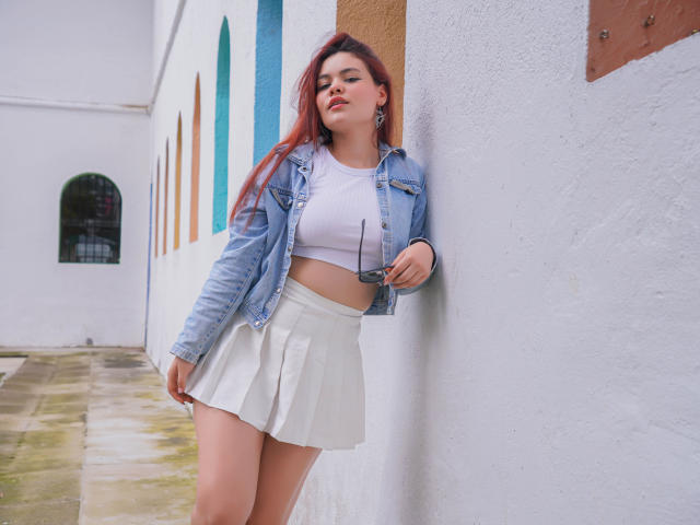 MiaPoe with red hair, white crop top, and white skirt, leaning against a wall in a denim jacket, holding sunglasses, showing her ass in a white pleated skirt.