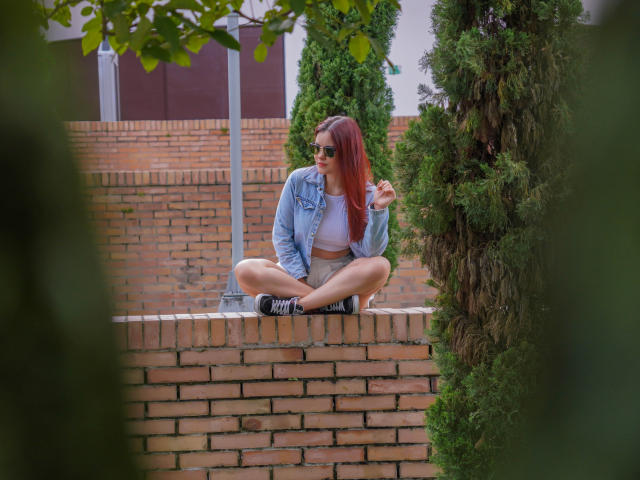 MiaPoe with red hair, wearing sunglasses, denim jacket, white crop top, and black shorts, sitting cross-legged on brick wall, touching her hair, surrounded by greenery.