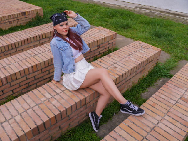 MiaPoe with long red hair, denim jacket, white skirt, black sneakers, and bandana, sitting on brick steps, showing her legs and ass, with green grass below.