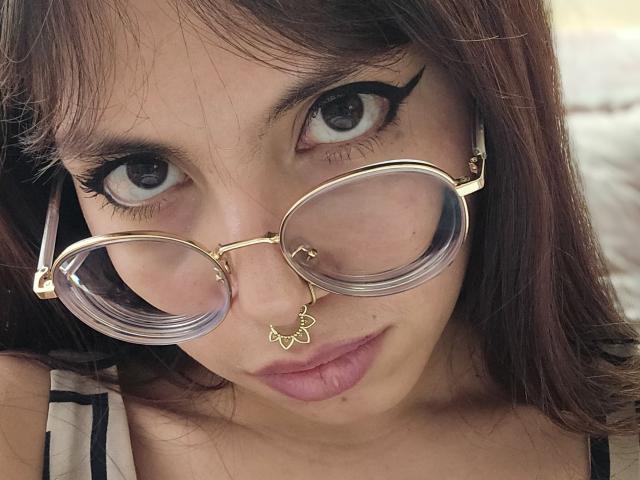 NaturalMariaHun with big brown eyes, wearing gold-rimmed glasses and a septum ring with a leaf design, has pink lipstick and dark brown hair, looking directly at the camera.