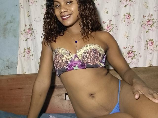 KennyReah has curly brown hair, smiling, wearing a strapless bra with purple and yellow floral patterns, and blue thong, sitting on a bed with a wooden headboard.