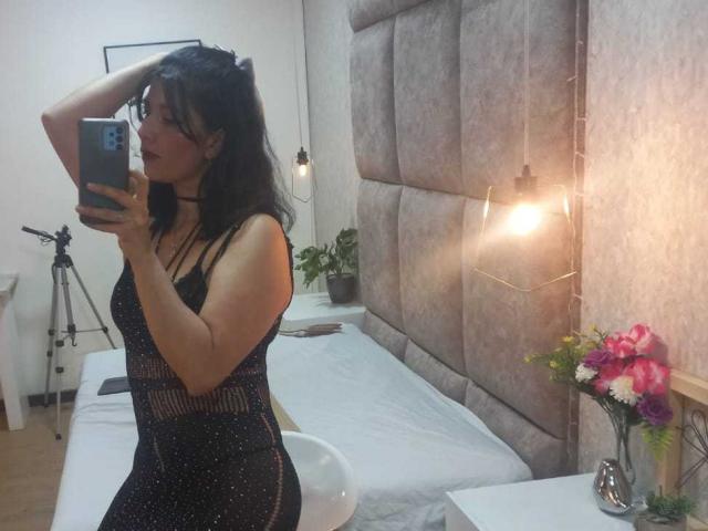 DianaMaris with dark hair, wearing a black, sparkly dress, taking a mirror selfie, showing her ass and small tits, standing by a bed with a tripod in the background.