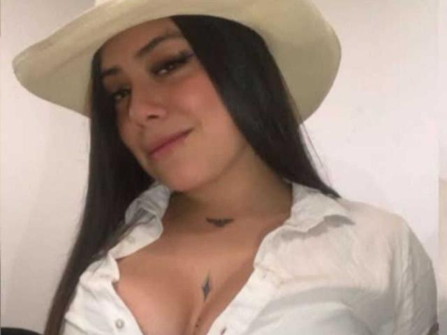 KatalinaSexy69 with long black hair, wearing a white cowboy hat and unbuttoned shirt, showing cleavage, has a small black tattoo above her tits.