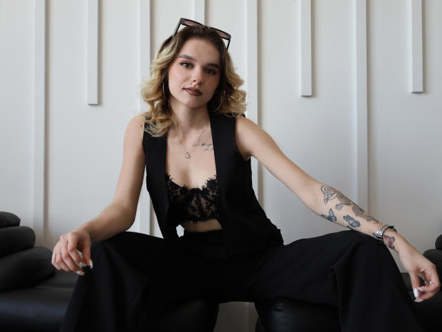 EmelyHailey with blonde curls, black lace bra, sleeveless black vest, and black pants, sitting with legs apart, showing tattoos on right arm, wearing black sunglasses on head.