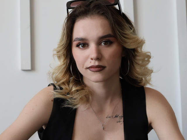 EmelyHailey, avec ses cheveux blonds bouclés, son haut noir sans manches, ses boucles d'oreilles, et ses lunettes de soleil sur la tête, regarde avec ses yeux marrons, un maquillage discret, un petit tatouage au-dessus de sa poitrine, et une expression neutre..