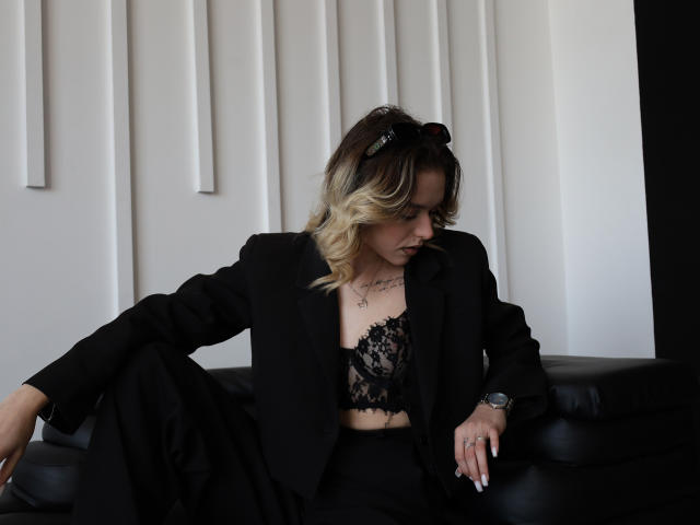 EmelyHailey mit blonden Ombre-Haaren, einem schwarzen Spitzen-BH und einem Blazer, sitzt auf einer schwarzen Ledersofa, blickt nach unten, trägt eine Sonnenbrille auf dem Kopf, Tattoos sind sichtbar, ein Ring am rechten Handgelenk..