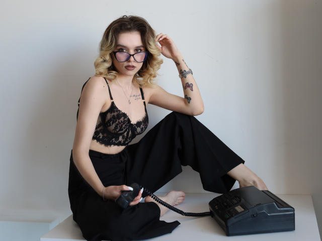 EmelyHailey, con i capelli biondi ricci, un reggiseno di pizzo nero, pantaloni neri, occhiali, tatuaggi, che tiene un telefono fisso, seduta su un pavimento bianco, con una mano tra i capelli, che guarda la fotocamera..