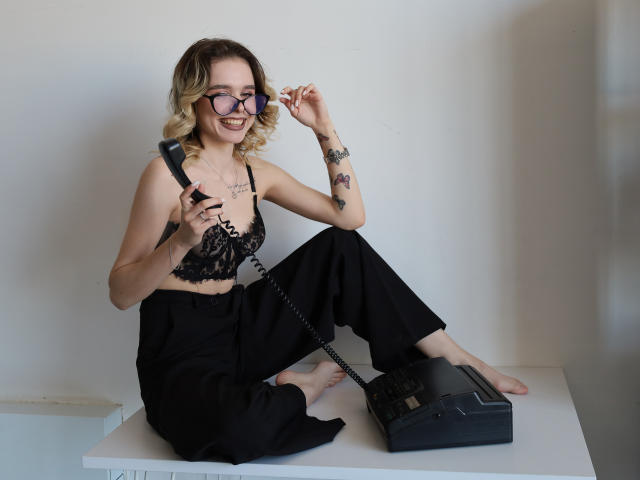EmelyHailey with curly blonde hair, glasses, black lace bra, black pants, holding old-school phone, sitting on white table, tattoo on left arm, smiling, barefoot, black phone on table.