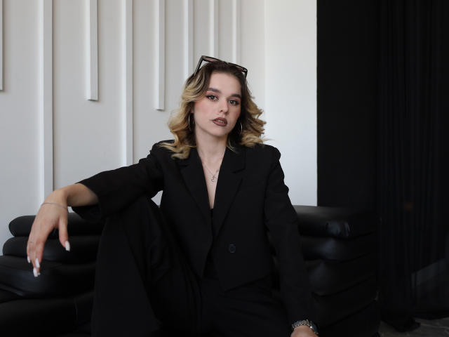 EmelyHailey with blonde curls, black suit, and dark lipstick, sitting on black leather couch, arm resting on knee, sunglasses on head, looking at camera, serious expression.