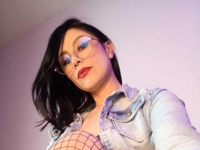 VioletaWhitee with glasses, red lipstick, black hair, denim jacket, fishnet top showing tits, slight smile, looking down at camera.