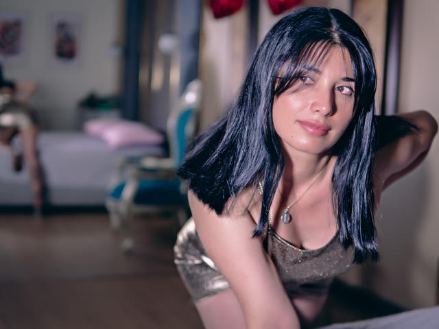 DianaMaris with black hair, wearing a shiny gold top, leaning forward with her hands behind her head, looking at the camera, showing her small tits and a subtle smile.