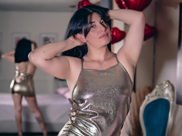 DianaMaris with black hair, wearing a shiny gold dress, arms raised, showing her tits, standing in a bedroom, looking at her reflection in a mirror.