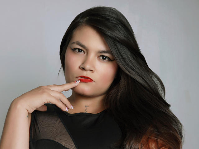 Camila Valera, con cabello negro largo, lápiz labial rojo y una expresión seria, toca su dedo contra sus labios rojos, vistiendo una blusa negra transparente que muestra un pequeño tatuaje en su cuello..