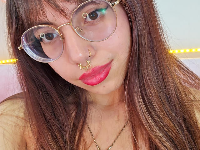NaturalMariaHun with brown hair, gold nose rings, and pink lipstick, wearing round glasses, smiling at the camera, showing off her red pout. Her tits are slightly visible.
