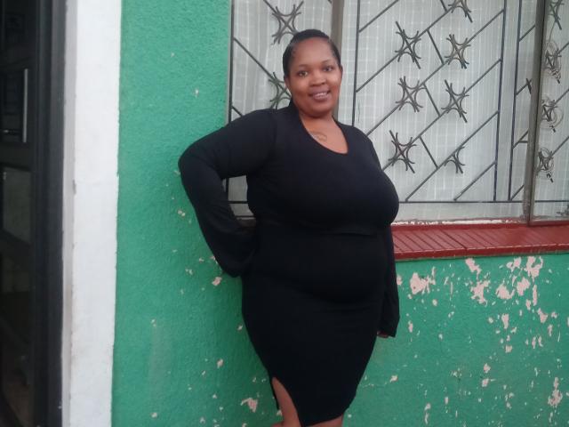 BiggyBoobs stands against a green wall, wearing a tight black dress that shows her ass and large boobs, with a confident smile, hands on hips, and short black hair.