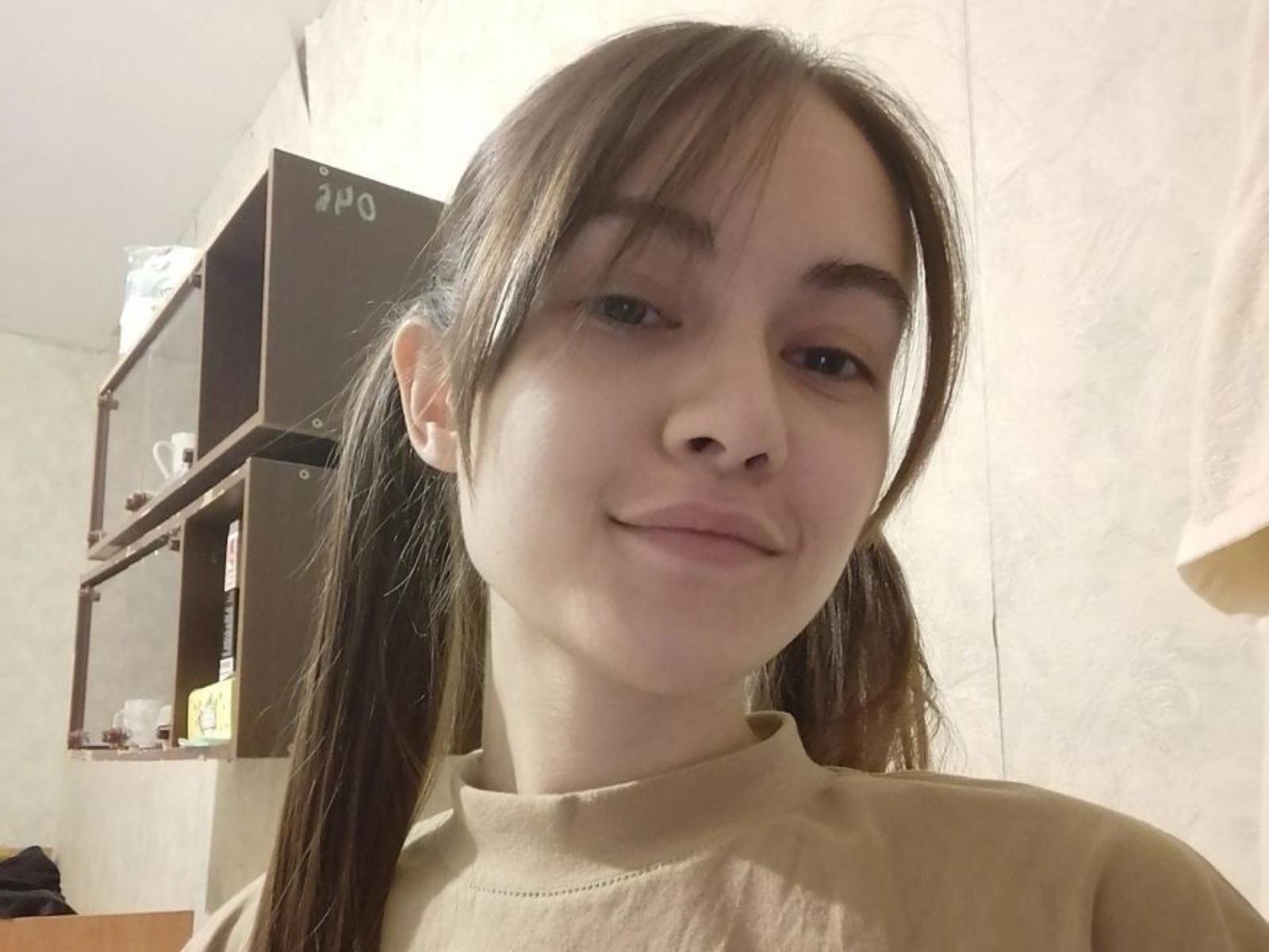 BeverlyBarrt with puffy brown tits, straight brown pigtails, and a light tan turtleneck, smiling slightly, showing pale skin and brown eyes in a softly lit room.