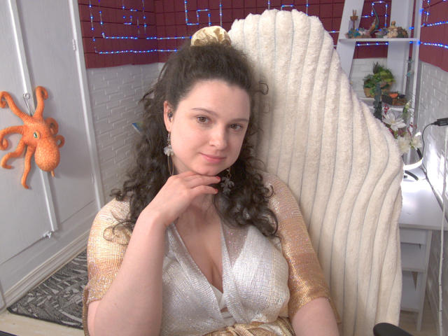 HandmadeMistress, with curly brown hair, wearing a sparkly gold and white top, sits in a ribbed white chair, touching her chin, smiling, with a cute orange octopus sticker.