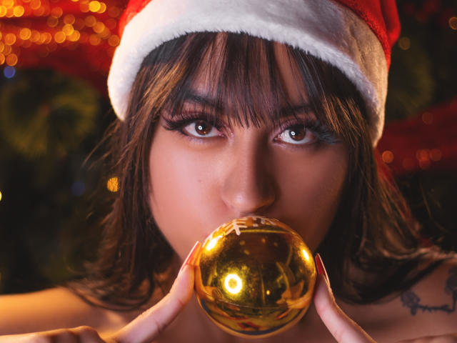 MadyJackson with dark brown hair, wearing a red and white Santa hat, biting a shiny gold ball, her brown eyes looking forward. Boob tattoo partially visible.