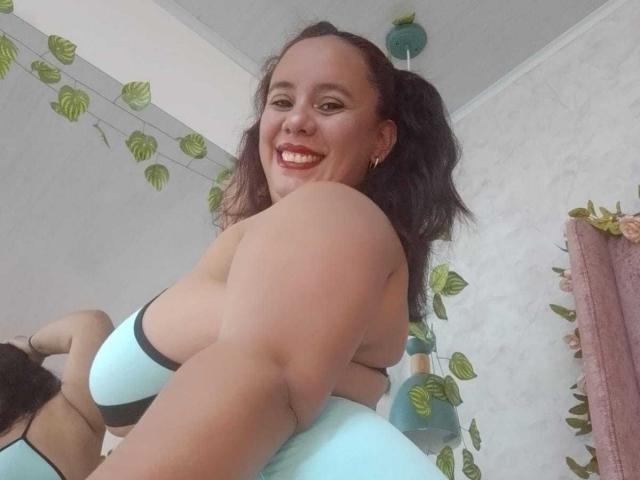 NicolletteDiaz with puffy ass, smiling, wearing a light blue bra with black straps, her brown hair in pigtails, reflecting in a mirror, showing her back and smiling face.