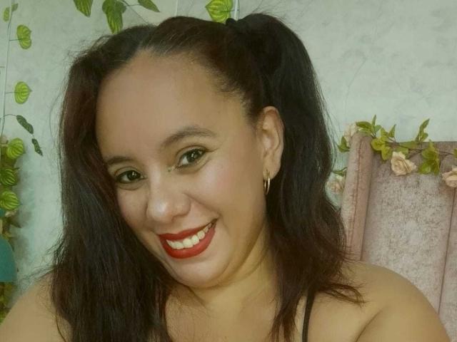 NicolletteDiaz has dark hair in pigtails, red lipstick, and a smile showing her teeth. She's wearing gold earrings and a black strap top. Her skin is light brown.