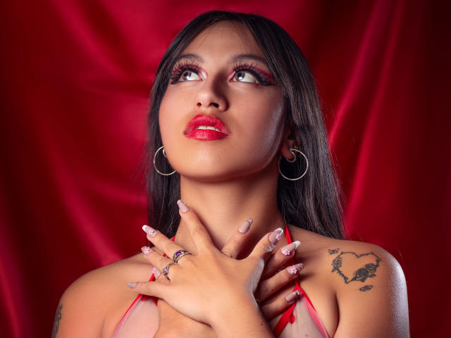 MadyJackson with red lipstick, long dark hair, hoop earrings, and tattoos on her shoulder, gazing upward, hands on her neck, wearing a red halter top, shiny nails.