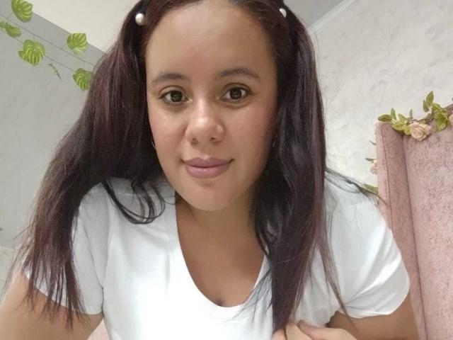 NicolletteDiaz with dark brown pigtails, wearing a white tee, has brown eyes, a nose ring, and a slight smile, showing her boobs in a selfie.