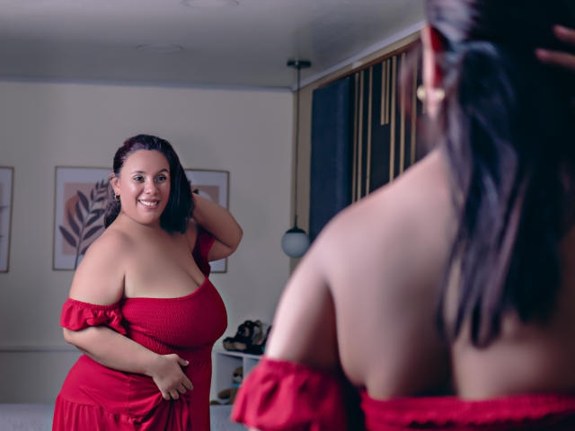 NicolletteDiaz with curvy, large tits in a red off-shoulder dress, smiling at another woman with her back to the camera, also in a red dress, in a room.