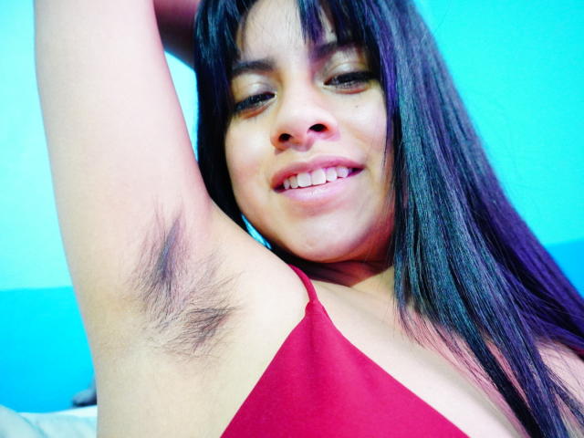 HairyDuchess with dark hair, smiling, raising arm to show armpit hair, wearing red top, blue background, and visible teeth.