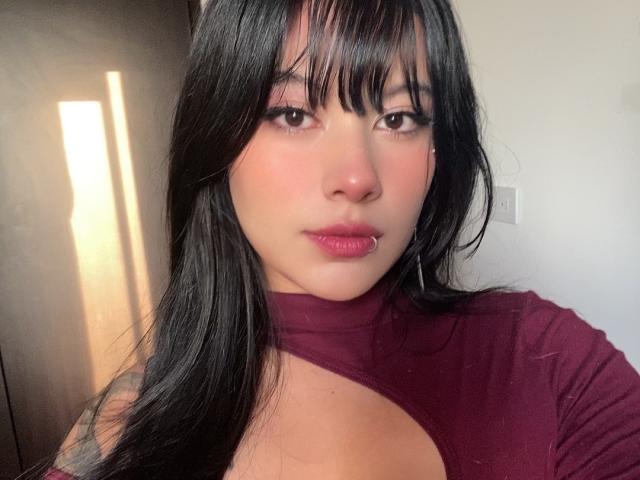 VelvetMoon with long black hair, bangs, and pink lipstick, wearing a maroon top with a cutout, has a slight tattoo on her left arm, and a cute nose.