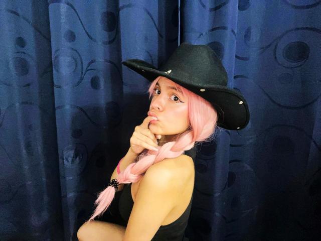 DanesaCronin with pink hair in a braid, wearing a black cowboy hat, black tank top, and sucking her finger, looking up with a cute expression.