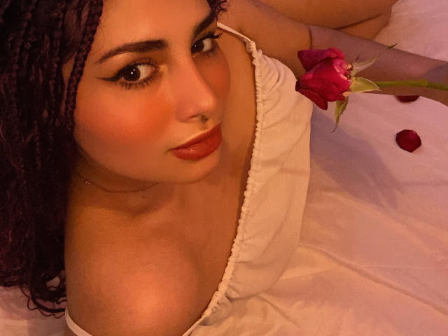 SweetSolei mit lila Zöpfen, rotem Lippenstift, weißem Oberteil mit Schulterauschnitt, die eine rote Rose hält, liegt auf einem weißen Bett, blickt leicht lächelnd nach oben, Nasenring, Rosenblätter..
