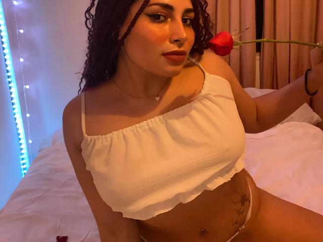 SweetSolei mit lockigem, braunem Haar, einem weißen Crop-Top, das ihre Brüste zeigt, hält eine rote Rose, hat gebräunte Haut, eine kleine Nasenring und ein Tattoo auf ihrer rechten Seite. Schlafzimmerszenario..