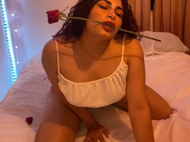 SweetSolei, met krullend donker haar, een neusring en gesloten ogen, bijt in een rozenstengel, draagt een witte crop top en knielt op een bed, waarbij haar borsten zichtbaar zijn, in een donkere kamer..