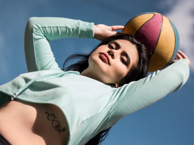 MaddieWillow sosteniendo una pelota, con los brazos levantados, vestida con una chaqueta verde menta con cremallera, que deja ver sus pechos y un tatuaje debajo del ombligo, con cabello oscuro y una expresión de labios fruncidos..