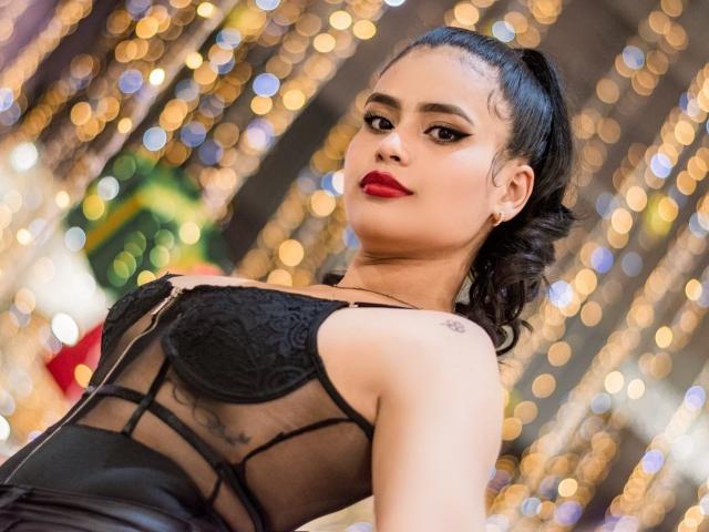 MaddieWillow, con i capelli scuri raccolti in una coda di cavallo, indossa un top di pizzo nero, rossetto rosso e piccoli orecchini, e guarda verso l'alto con un leggero sorriso. Sfondo con effetto bokeh..