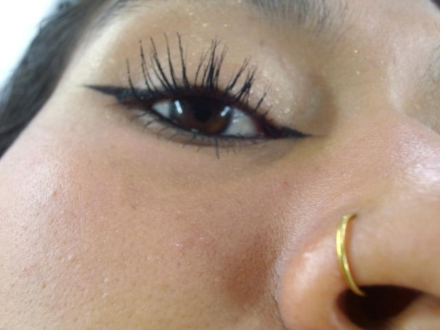 AnnieDarling's face with a gold nose ring, brown eye, long black eyelashes, and shimmery eyeshadow. Her skin is slightly blemished with a soft, warm tone.