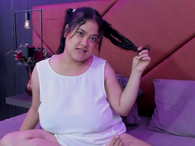 JuliaHill, chubby with small tits, sits on a bed, holding her black braid, wearing a white sleeveless top, and a white skirt, with a pink headboard in the background.